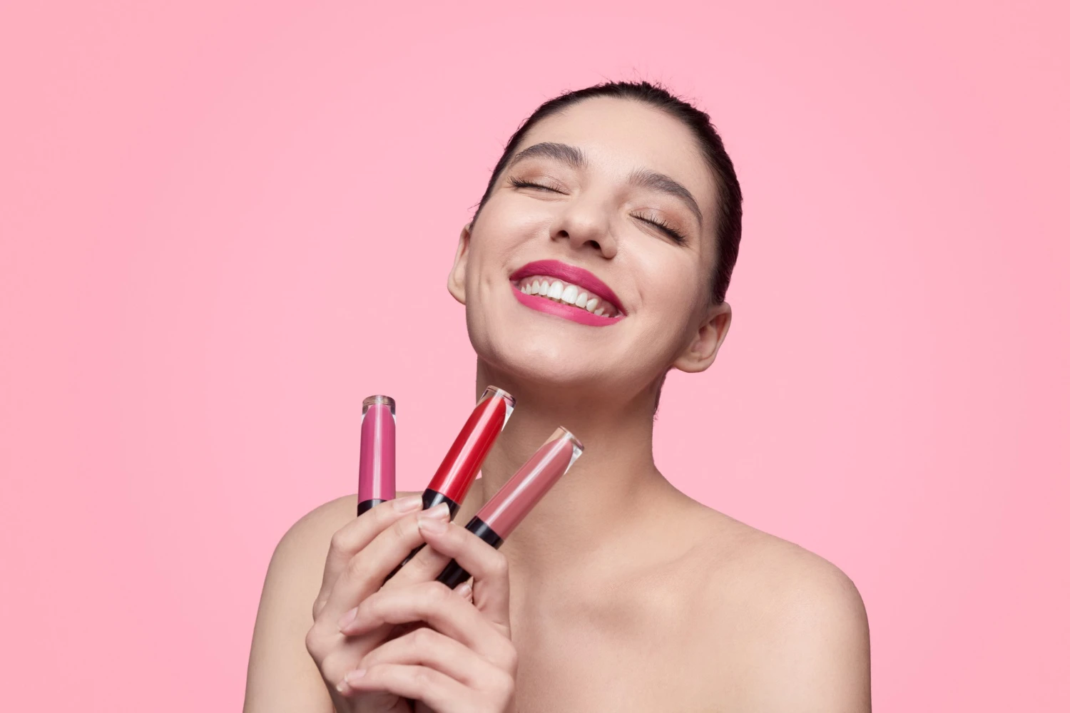 Mulher sorridente com maquiagem, segurando e mostrando batom cintiliante e batom em tons de rosa e vermelho em fundo rosa liso, com pele iluminada.
