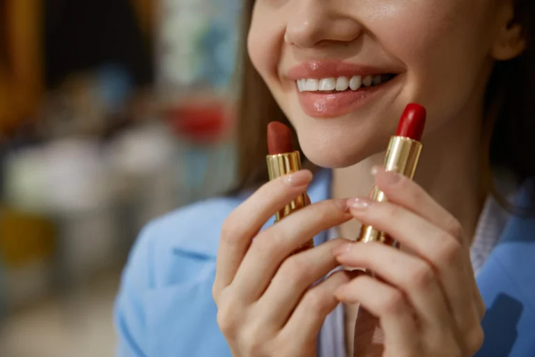 close no rosto sorridente de uma mulher aplicando batom cintiliante vermelho em um estojo dourado, com mãos levantadas e fundo desfocado