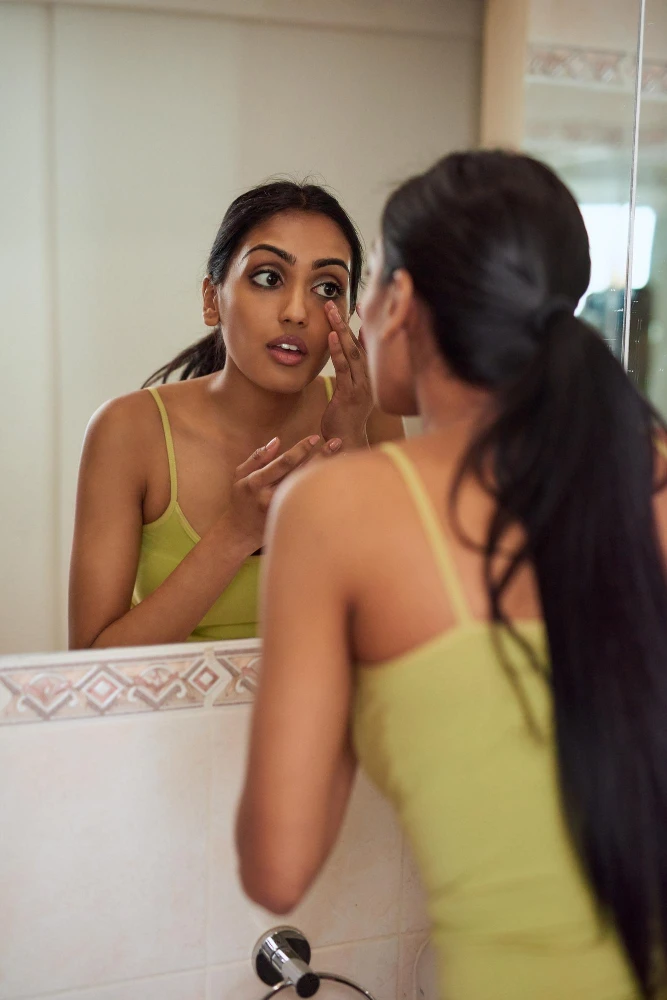 Mulher em frente ao espelho no banheiro, aplicando cuidado facial com as mãos, enquanto observa o próprio reflexo; imagem remete a rotina de pele mista