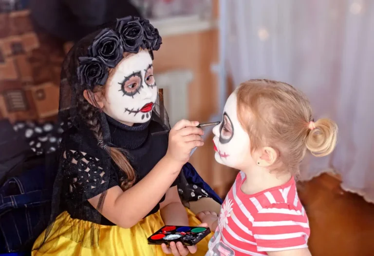 halloween e dia das crianças: Criança brinca com fantasia de Halloween ao lado de um adulto mascarado com maquiagem de caveira, segurando um pincel e aplicando tinta no rosto em comemoração ao dia das crianças.