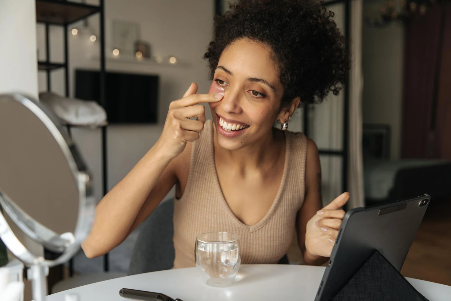 Mulher sorridente aplicando maquiagem no rosto diante do espelho, fazendo maquiagem dia a dia com look natural em ambiente de sala com mesa e celular