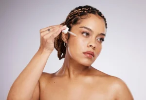 Mulher aplicando serum facial com conta-gotas no rosto, com expressão natural e iluminação suave em fundo claro, mostrando cuidado com a pele