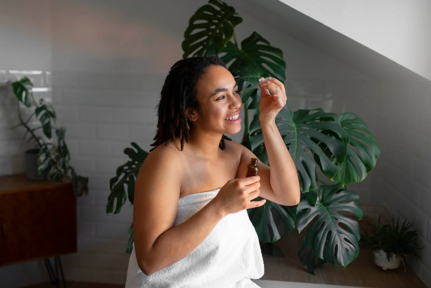 Mulher sorrindo no banheiro com iluminação natural, segurando um frasco de serum facial e aplicando no rosto, ao lado de plantas decorativas e fundo com azulejos claros.