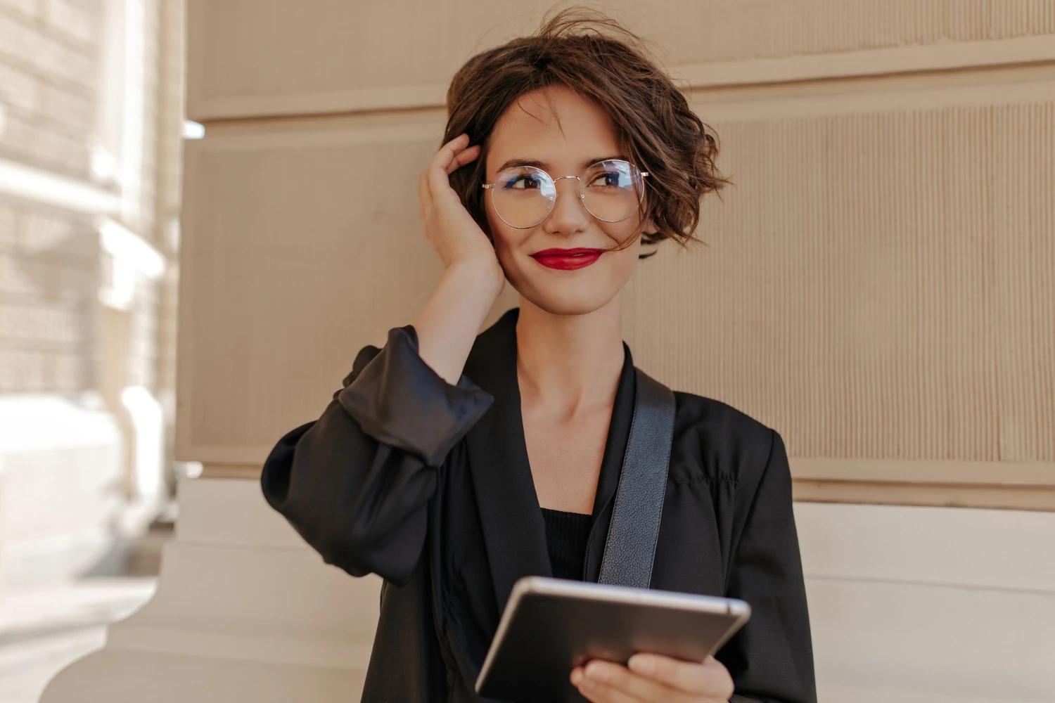 Mulher usando óculos com armação azul e batom vermelho, em ambiente interno, segurando um celular e ajustando o cabelo; maquiagem dia a dia em estilo moderno.