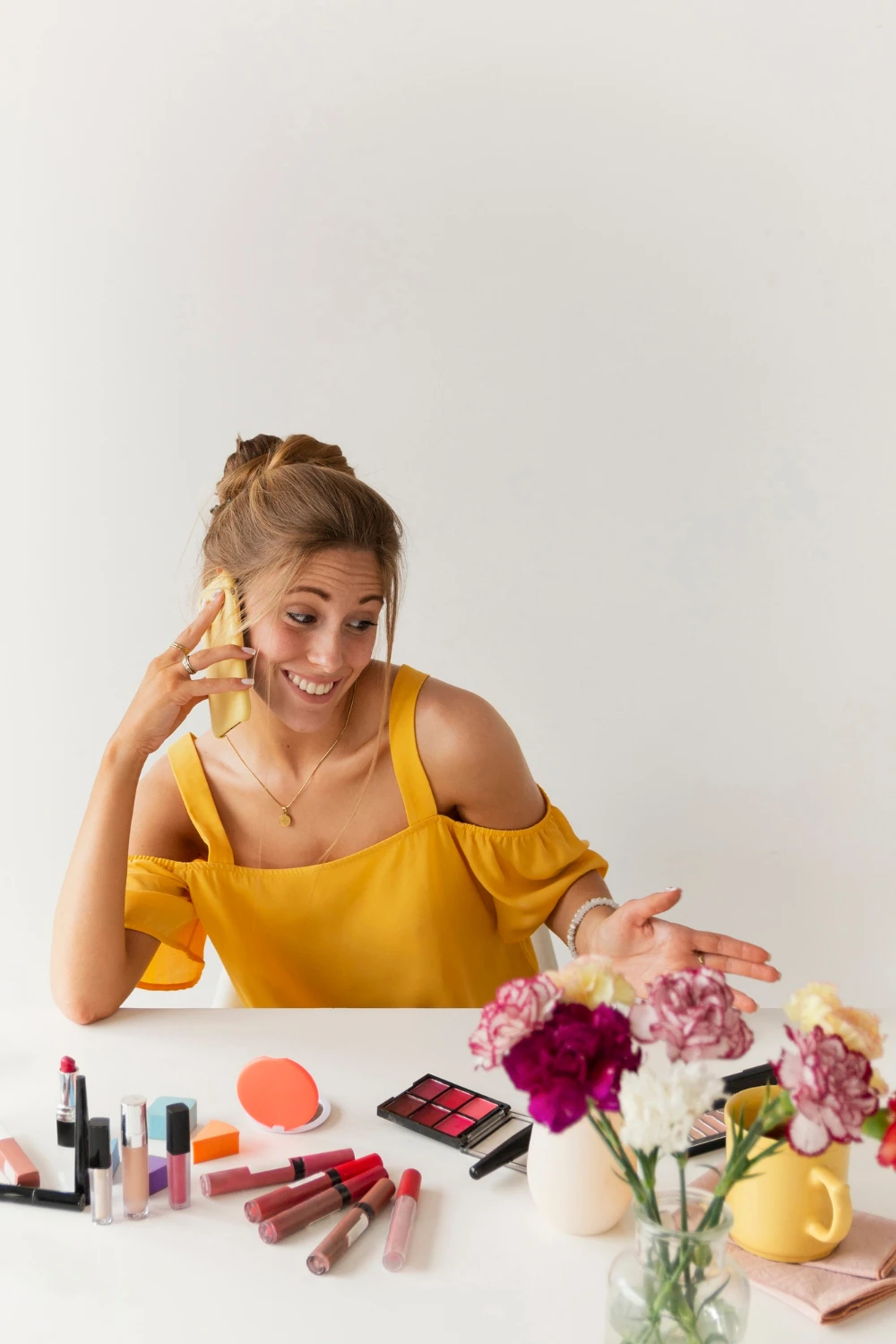 o que vender na primavera: Mulher sorridente em mesa de maquiagem com produtos cosméticos, flores e um celular ao fundo, usando blusa amarela e compondo um look de primavera