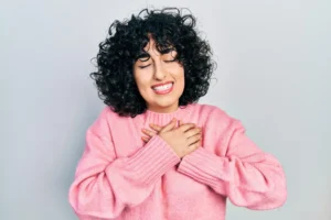 Outubro rosa: mulher com cabelo cacheado usando suéter rosa, sorrindo e com as mãos no peito em sinal de conscientização