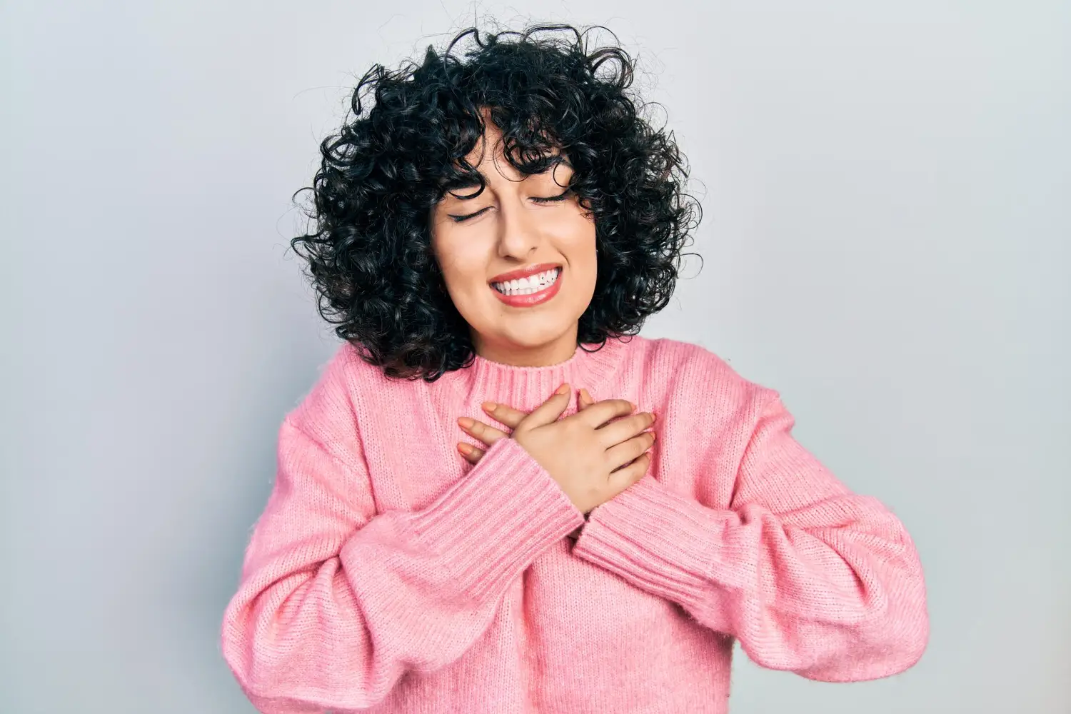 Outubro rosa: mulher com cabelo cacheado usando suéter rosa, sorrindo e com as mãos no peito em sinal de conscientização