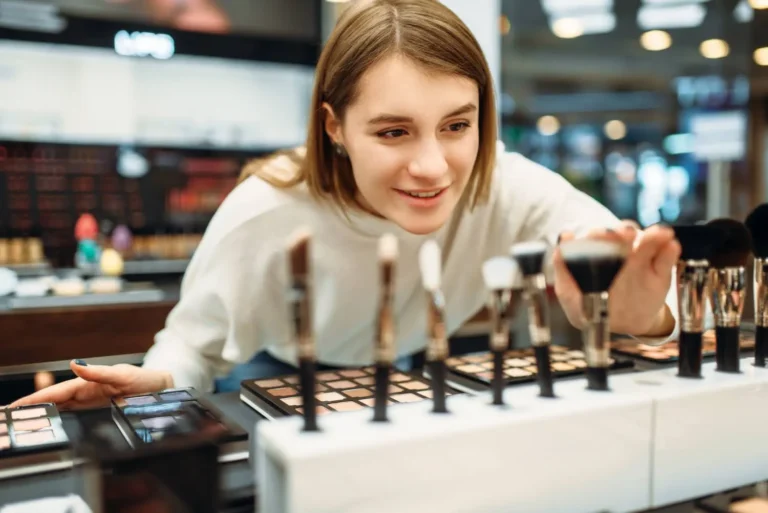 black friday: Vendedora em loja de cosméticos apresenta produtos de maquiagem em balcão, com pincéis e paleta de sombras ao fundo, em ambiente iluminado e organizado.